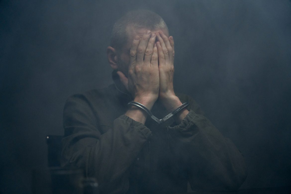 Close-up of a suspect in handcuffs closing his face with hands in a dark fogged room Spells to free prisoner