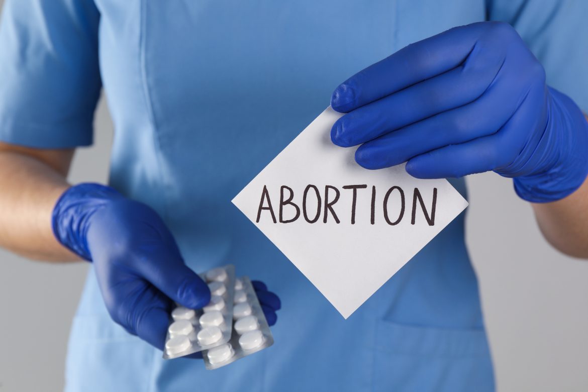 Doctor holding pills and paper card with word ABORTION on light grey background, closeup Power of command